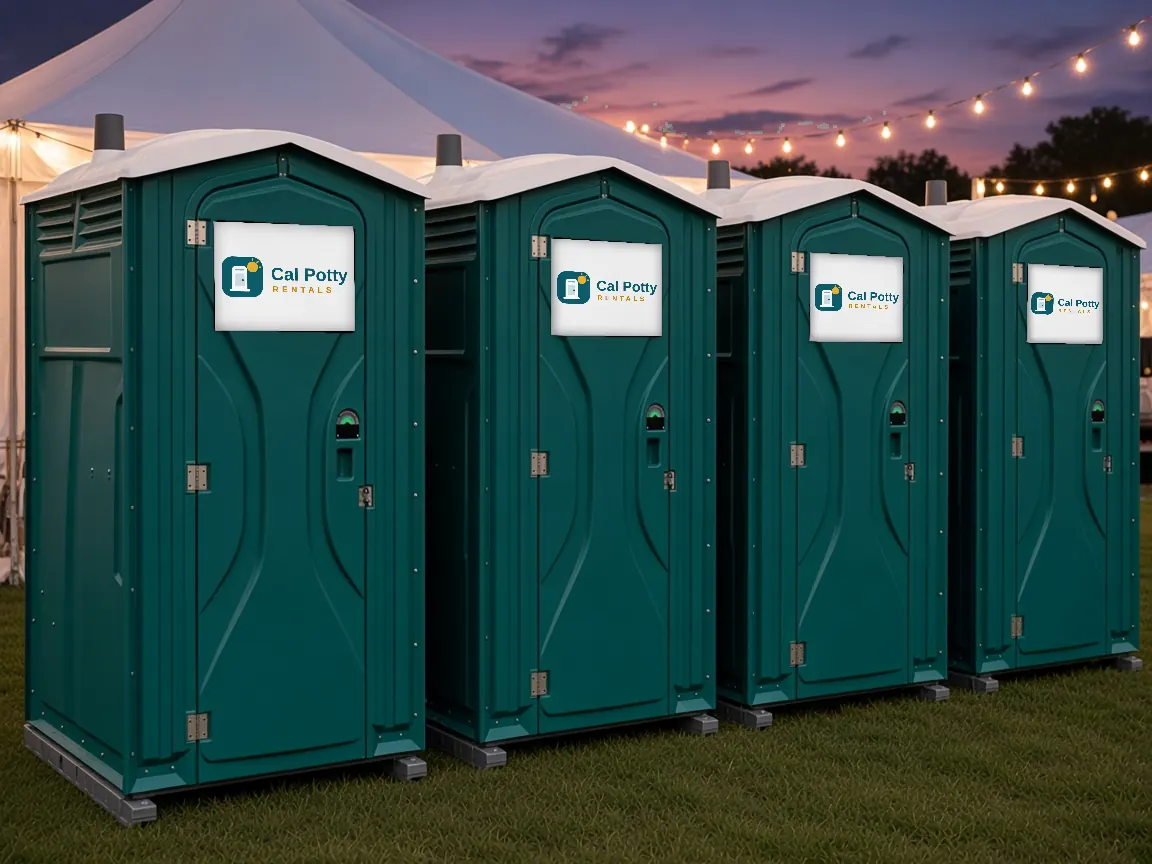 Four teal portable toilets on a lawn at dusk under string lights