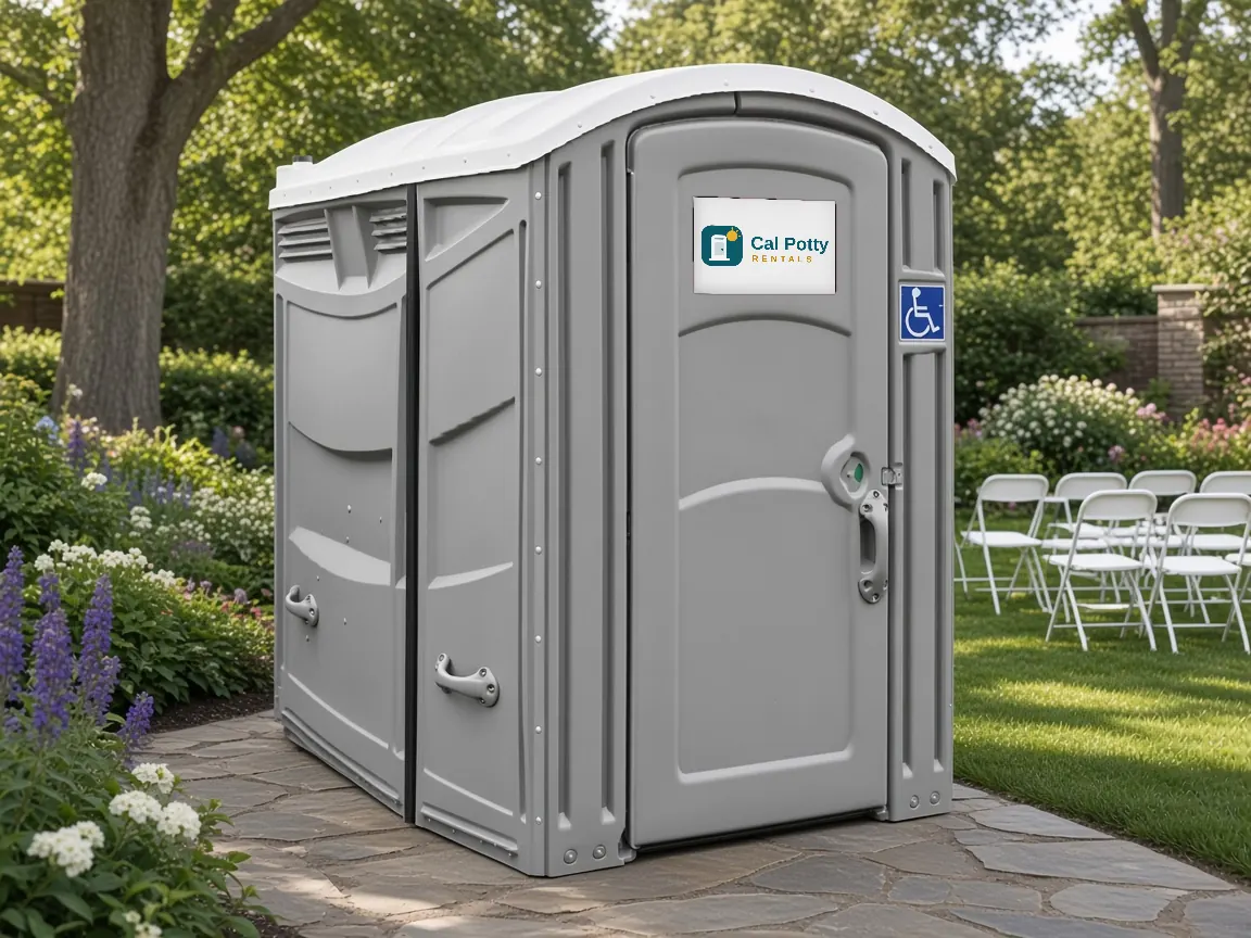 A grey ADA-accessible portable restroom with a wheelchair sign on a garden path near lawn chairs